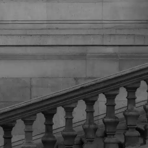 Schwarz-weißes Treppengeländer mit wiederkehrenden Säulen in diagonaler Perspektive, symbolisiert strukturierte soziale Bewegung und Reproduktion.